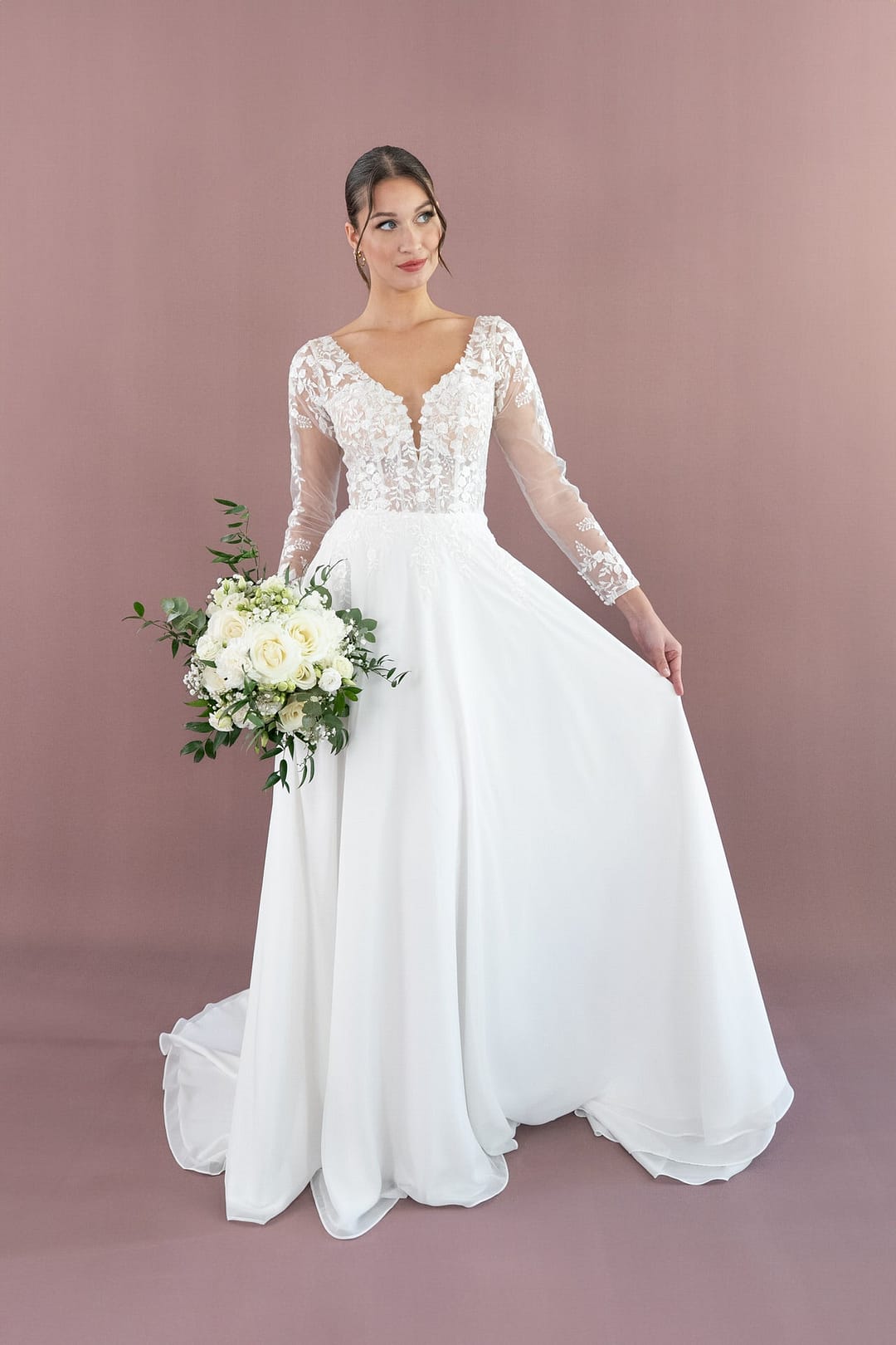 V26-A08_Web_1 Eine Braut im langärmeligen Bridonista V26-A08 Brautkleid hält einen Strauß weißer Blumen und Grünzeug, steht vor einem unifarbenen mauvefarbenen Hintergrund und hebt mit einer Hand sanft ihren Rock.