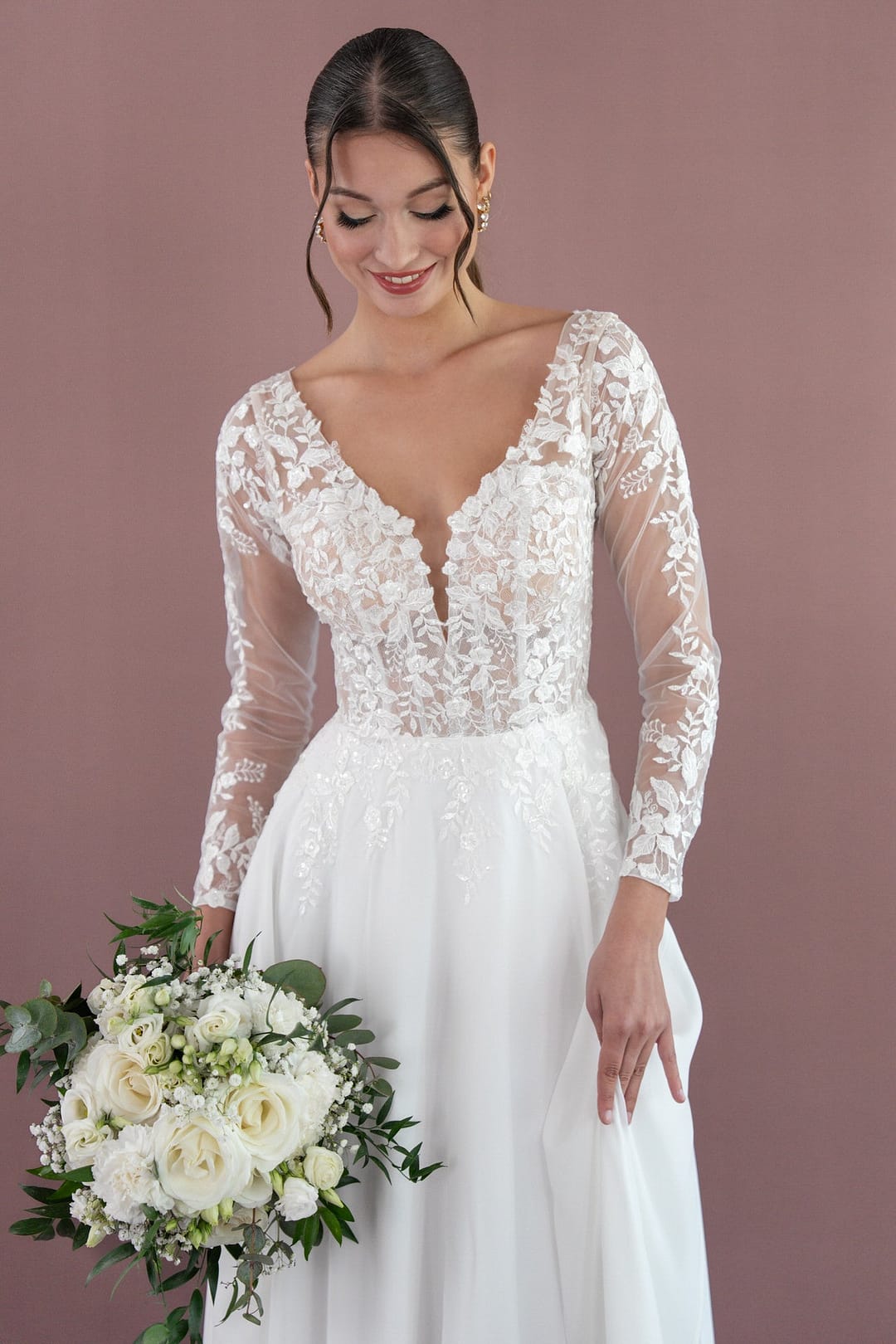 V26-A08_Web_3 Eine lächelnde Braut trägt das Bridonista V26-A08, ein weißes, langärmeliges Hochzeitskleid, und hält einen Strauß weißer Rosen und Grünpflanzen vor einem einfarbig lilafarbenen Hintergrund.