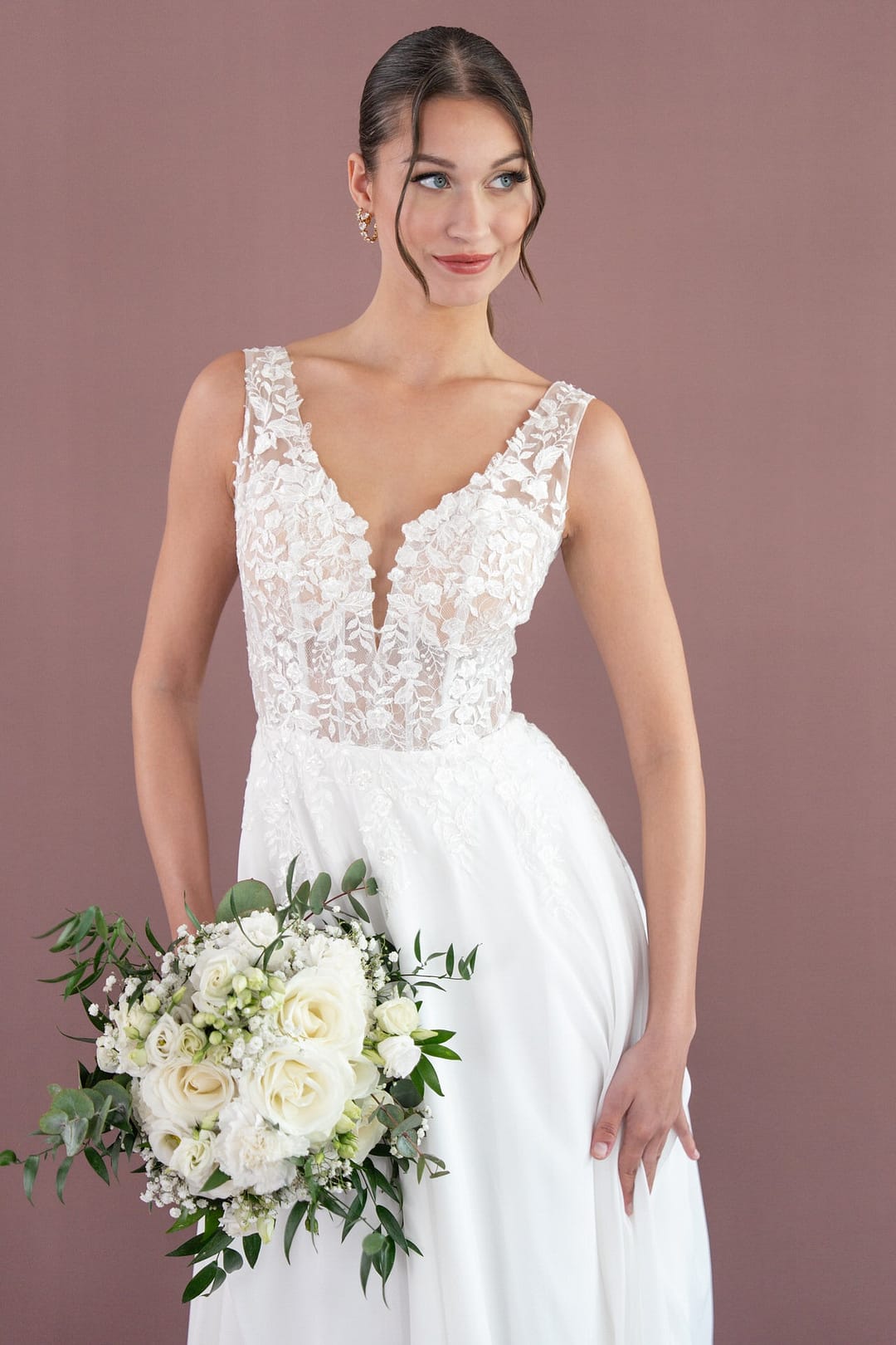 V26-A08_Web_4 Eine Braut im ärmellosen Bridonista V26-A08 Brautkleid mit tiefem V-Ausschnitt hält einen Strauß weißer Rosen und Grünzeug, steht vor einem unifarbenen mauvefarbenen Hintergrund und lächelt sanft.