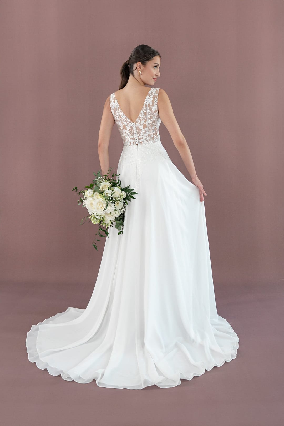 V26-A08_Web_5 Eine Braut trägt das Bridonista V26-A08, ein weißes Kleid mit Spitzenmieder und fließendem Rock. Sie posiert vor einem lilafarbenen Hintergrund mit einem Blumenstrauß und zeigt den tiefen Rücken und die Schleppe des Kleides, während sie über ihre Schulter blickt.