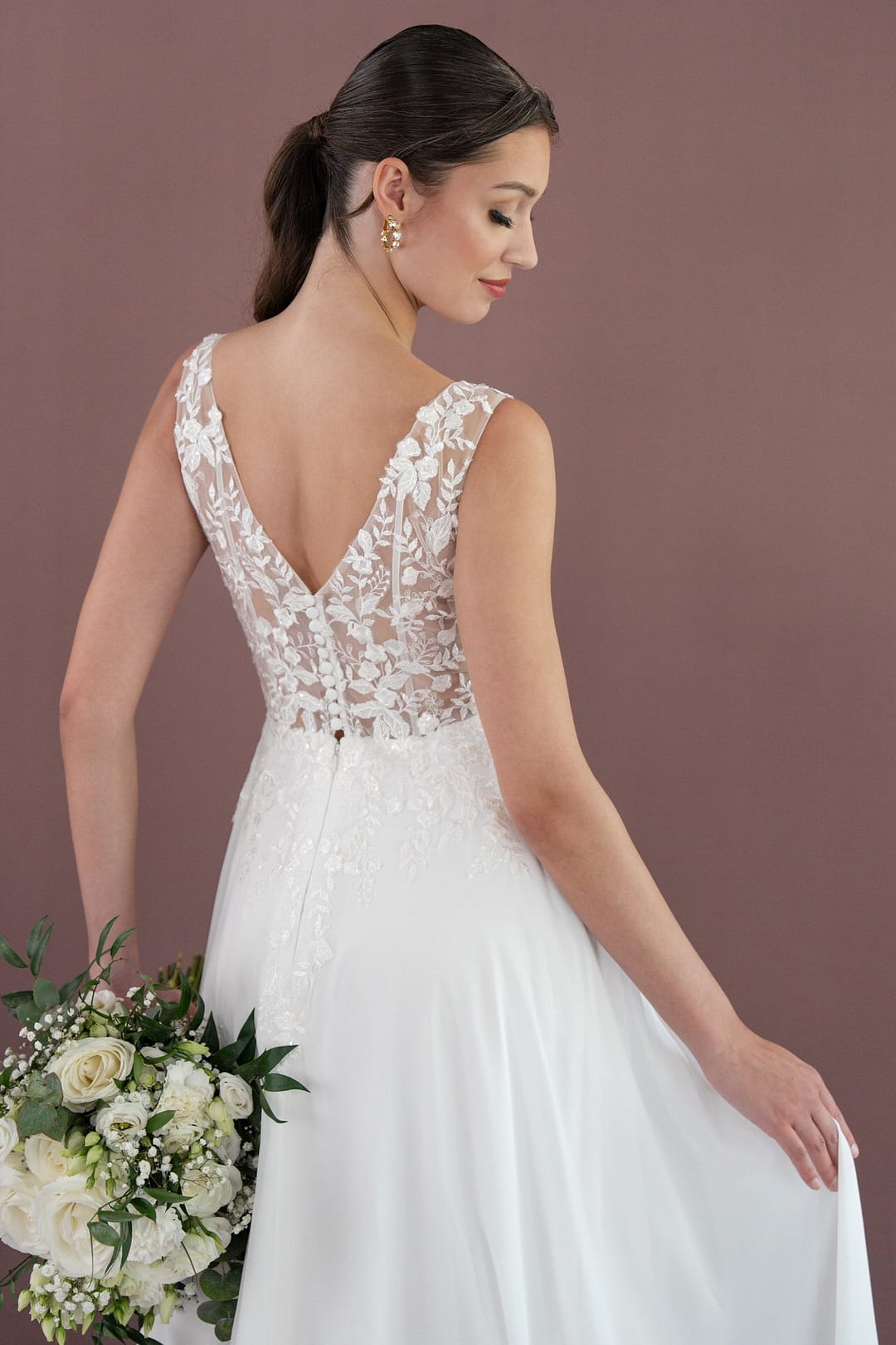 V26-A08_Web_6 Eine Braut in dem ärmellosen Spitzenkleid Bridonista V26-A08 hält einen Strauß weißer Blumen und Grünzeug und wendet sich ab, um die aufwendigen Spitzen- und Knopfdetails zu präsentieren. Ihr Haar ist zu einem niedrigen Pferdeschwanz gebunden und steht vor einem schlichten Hintergrund.