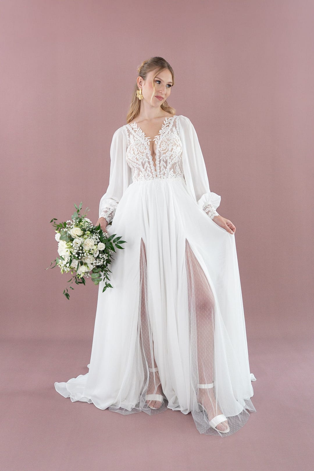 V26-A09_Web_1 Eine Frau trägt das langärmelige Bridonista V26-A09 Hochzeitskleid mit Spitzen-Details und zwei vorderen Schlitzen und hält einen Strauß weißer und grüner Blumen vor rosa Hintergrund.