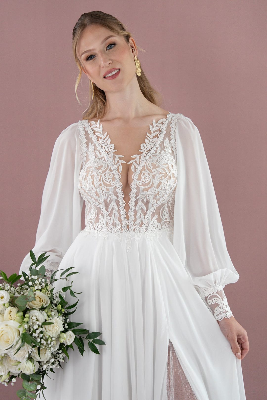 V26-A09_Web_2 Eine Frau im Bridonista V26-A09, einem weißen Spitzenhochzeitskleid mit langen durchsichtigen Ärmeln, hält einen Strauß weißer Rosen und Grünzeug in der Hand und lächelt sanft in die Kamera vor einem zart lila Hintergrund.