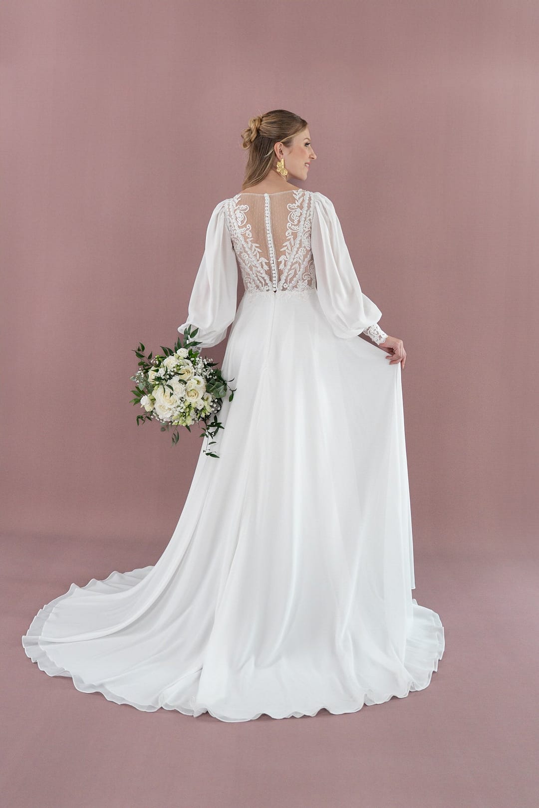 V26-A09_Web_4 Eine Braut, die einen Blumenstrauß in der Hand hält, blickt von sich weg und präsentiert das Bridonista V26-A09 - ein weißes Kleid mit durchsichtigem Spitzenrücken, langen Puffärmeln und einer fließenden Schleppe - vor einem Hintergrund in zartem Lila.