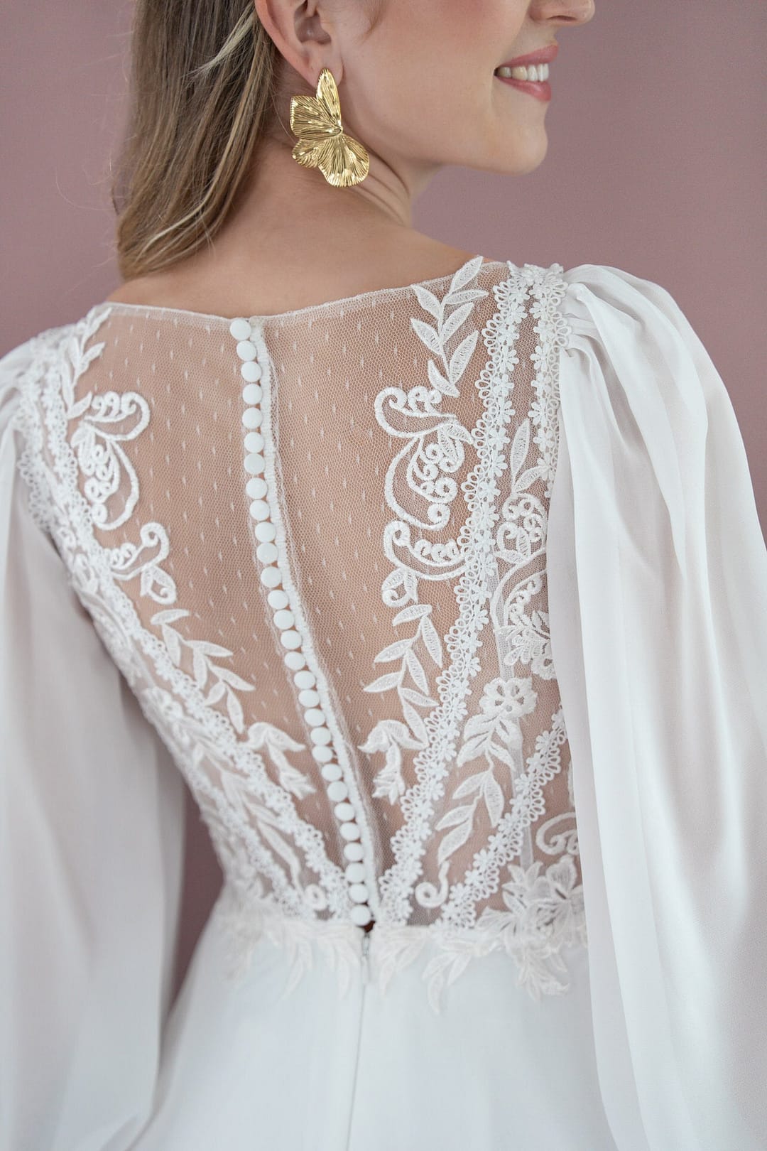 V26-A09_Web_5 Eine Frau trägt das Bridonista V26-A09, ein weißes Brautkleid mit durchsichtiger Spitze und stoffbezogenen Knöpfen am Rücken. Sie kombiniert es mit goldenen Schmetterlingsohrringen und lächelt, von der Seite auf einem zartrosa Hintergrund gezeigt - eine ideale Brautmoden-Inspiration.