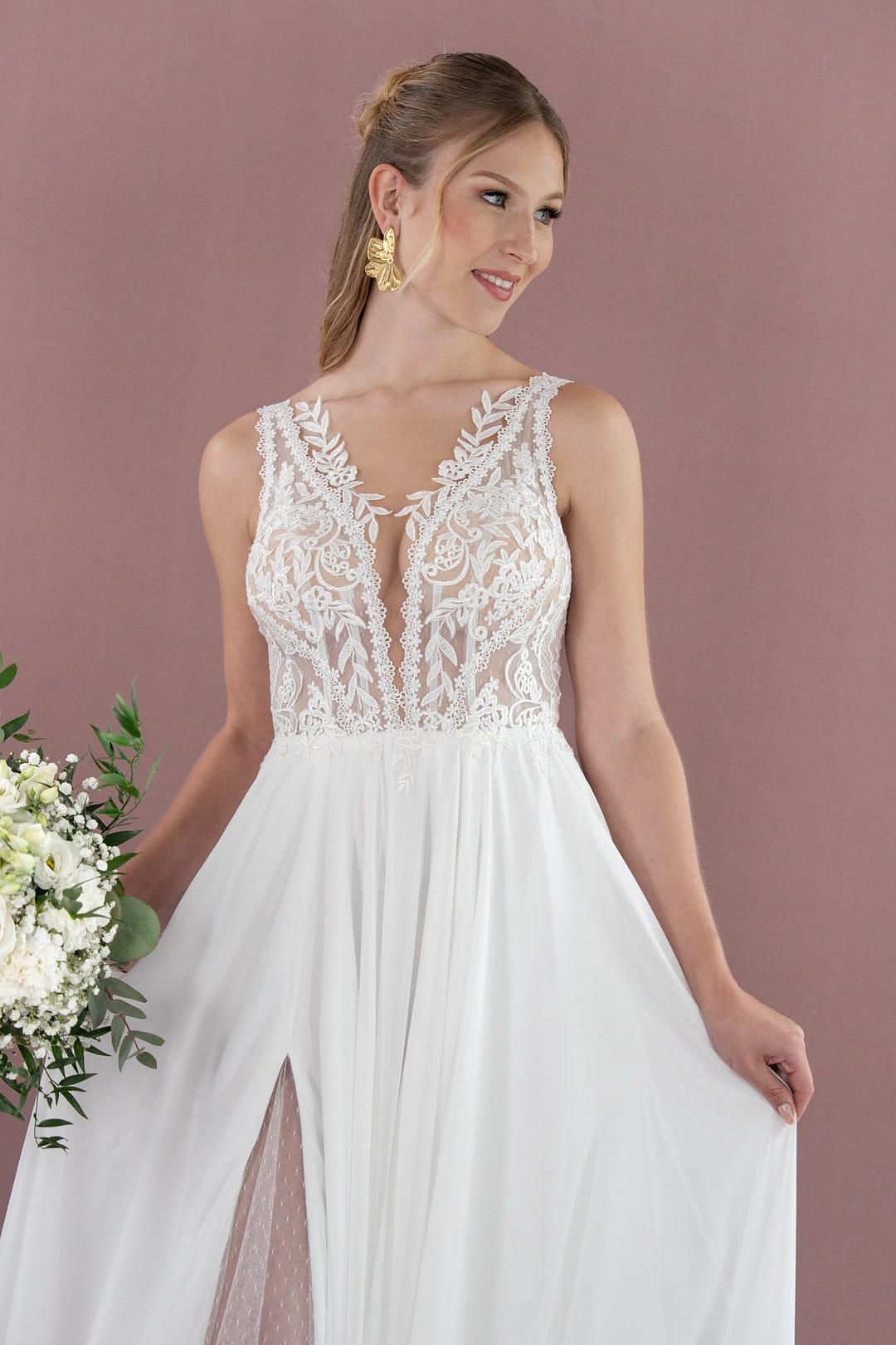 V26-A09_Web_6 Eine Frau in dem weißen Spitzenbrautkleid Bridonista V26-A09 mit durchsichtigem, besticktem Mieder und einem fließenden Rock mit Schlitz hält weiße Blumen. Ihr hellbraunes Haar ist zurückgestylt, während sie vor einem malvenfarbenen Hintergrund posiert.