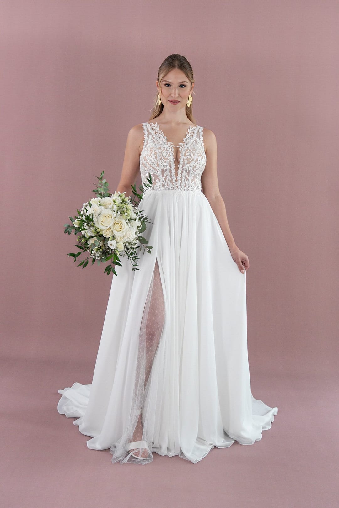 V26-A09_Web_7 Eine Frau, die das Bridonista V26-A09, ein weißes Spitzenhochzeitskleid mit durchsichtigem Mieder und hohem Schlitz, trägt, steht vor einem rosafarbenen Hintergrund und hält einen Strauß aus weißen und cremefarbenen Blumen mit Grünzeug.
