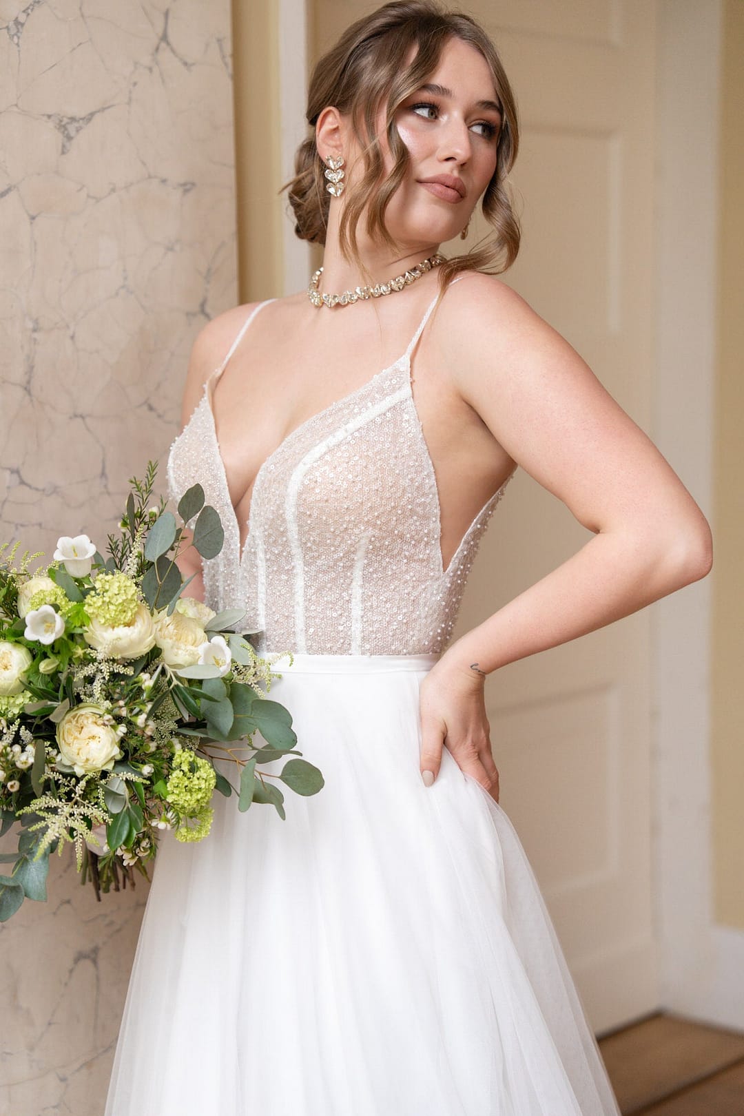 V26-A23_Web_1 Eine Braut steht in Bridonista V26-A23, einem ärmellosen Spitzenhochzeitskleid mit durchscheinendem Mieder und Tüllrock, und hält einen Strauß weißer und grüner Blumen. Ihre gewellte Hochsteckfrisur und ihr eleganter Schmuck vervollständigen den Look.