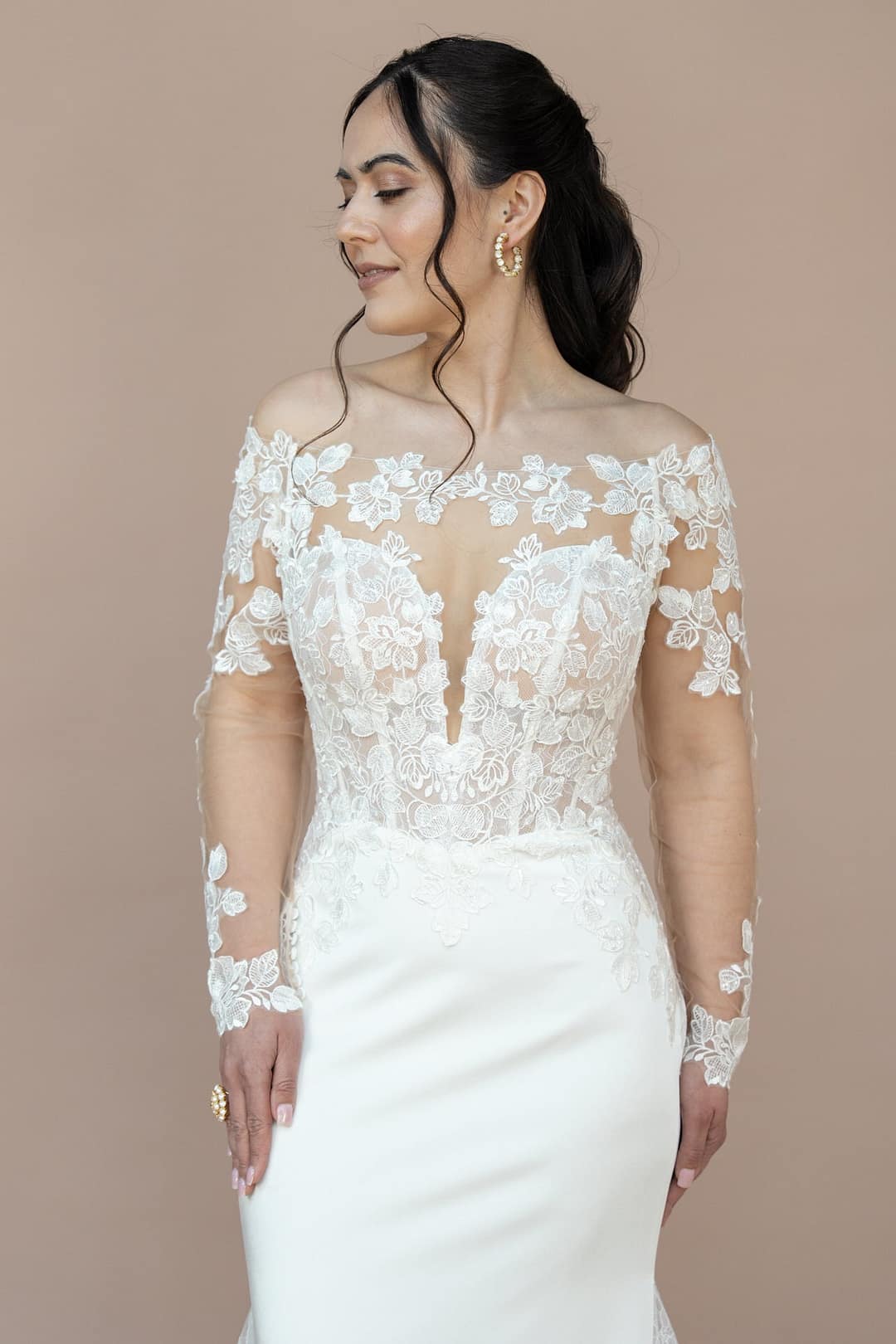 V26-F05_Web_2 Eine Frau in dem schulterfreien, langärmeligen weißen Brautkleid Bridonista V26-F05 mit floraler Spitze steht vor einem beigen Hintergrund und blickt mit einem sanften Lächeln zur Seite - zeitlose Brautmoden-Eleganz.