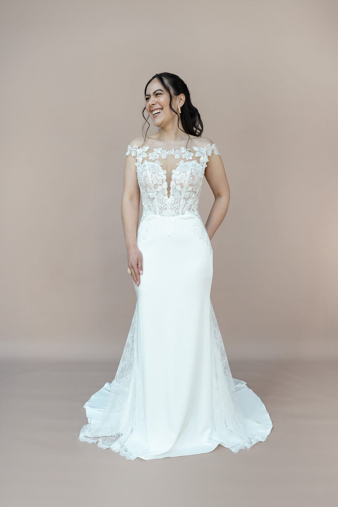 V26-F05_Web_3 Eine lächelnde Frau trägt das Bridonista V26-F05, ein bodenlanges Brautkleid mit Spitzenmieder und Flügelärmeln, vor einem schlichten hellbeigen Hintergrund. Ihr sanft gewelltes Haar unterstreicht den eleganten Brautmoden.