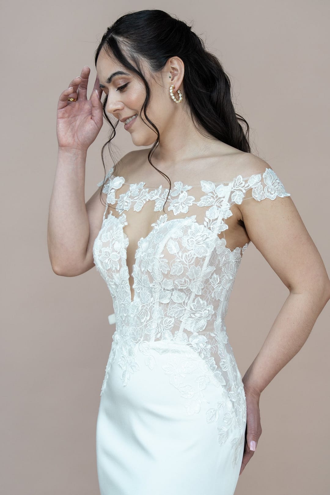 V26-F05_Web_4 Eine Frau in einem weißen schulterfreien Bridonista V26-F05-Brautkleid aus Spitze lächelt und berührt ihr Gesicht. Sie hat langes, gewelltes Haar und goldene Ohrringe und steht vor einem schlichten hellbraunen Hintergrund.