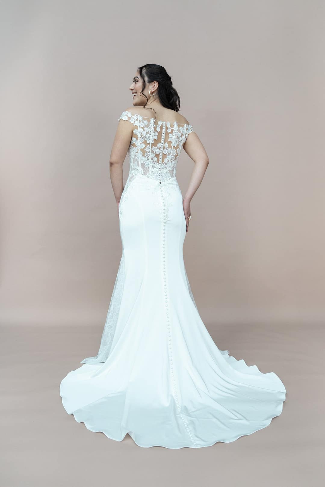 V26-F05_Web_5 Eine Frau in dem weißen Brautkleid V26-F05 von Bridonista mit langer Schleppe steht mit dem Rücken zugewandt und präsentiert die aufwendige Spitze und die Knöpfe vor einem einfarbigen beigen Hintergrund.