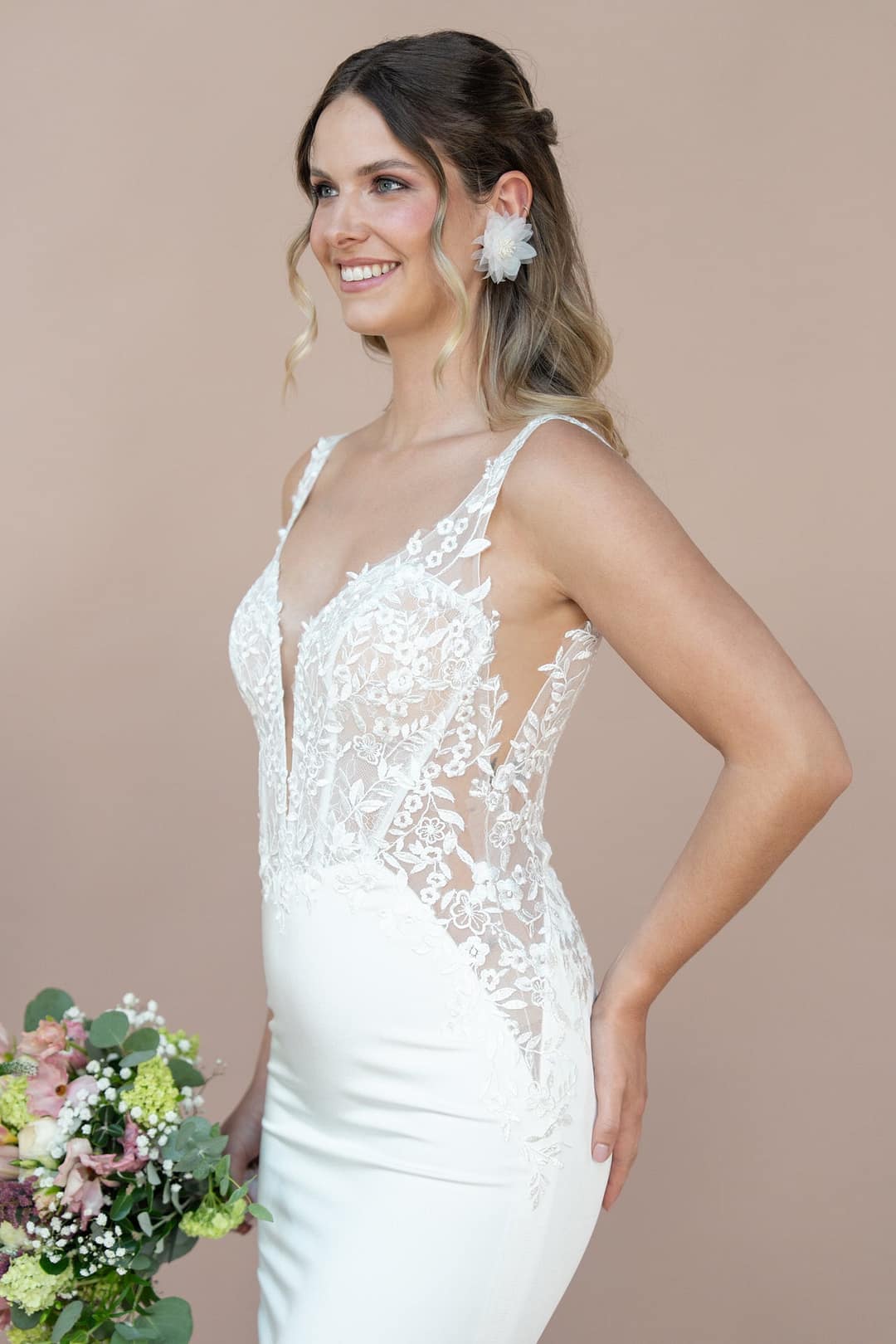 V26-F07_Web_3 Eine Frau im weißen, spitzenverzierten Bridonista V26-F07 Brautkleid hält einen Blumenstrauß, lächelt mit gewelltem Haar und einem floralen Accessoire vor einem schlichten beigen Hintergrund - die perfekte Inspiration für Brautmoden.
