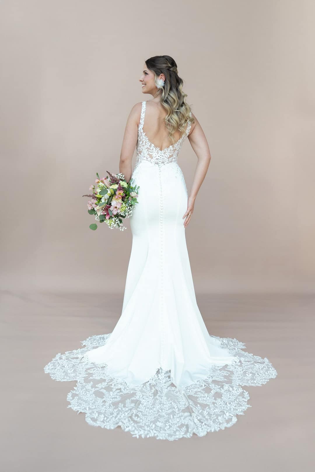 V26-F07_Web_5 Eine Frau in Bridonista V26-F07, einem taillierten weißen Spitzenbrautkleid mit langer Schleppe, steht lächelnd mit einem bunten Blumenstrauß in der Hand vor einem beigen Hintergrund - die perfekte Inspiration für Brautmoden-Fans.