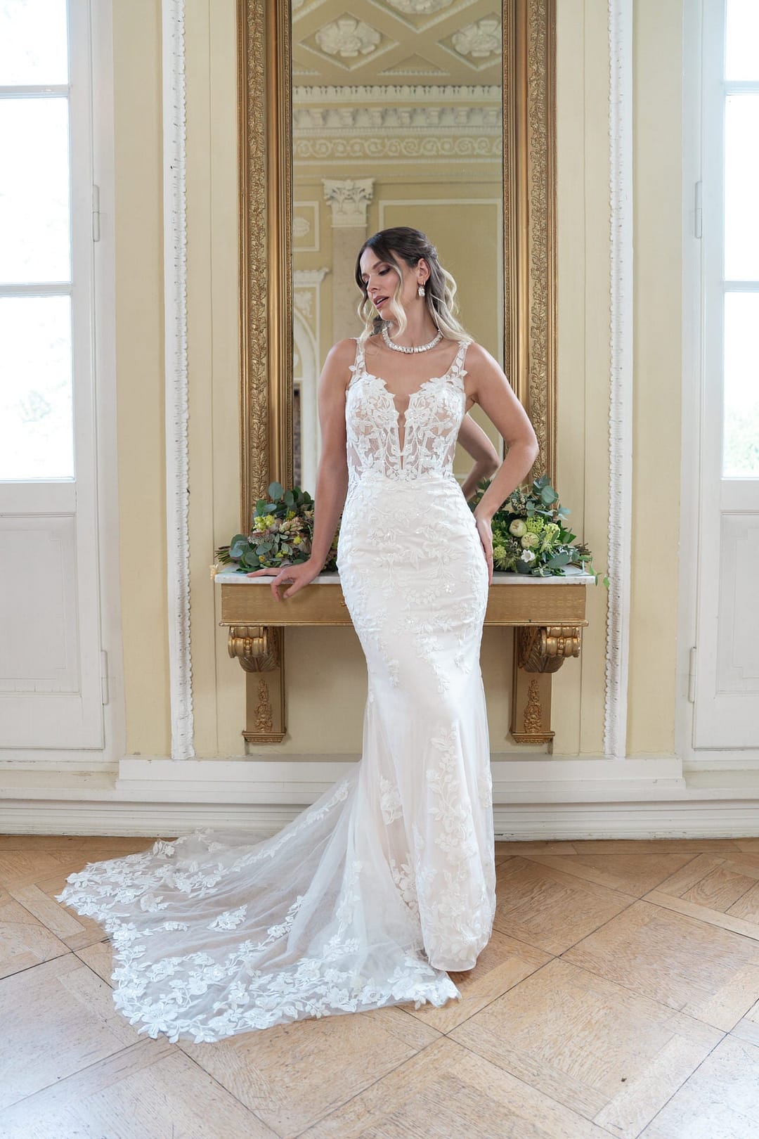 V26-F13_Web_1 Eine Frau, die das Bridonista V26-F13, ein weißes Spitzenbrautkleid mit langer Schleppe, trägt, lächelt vor einem goldgerahmten Spiegel und einer Blumenauslage in einem eleganten Raum mit hohen Fenstern und kunstvollem Dekor.