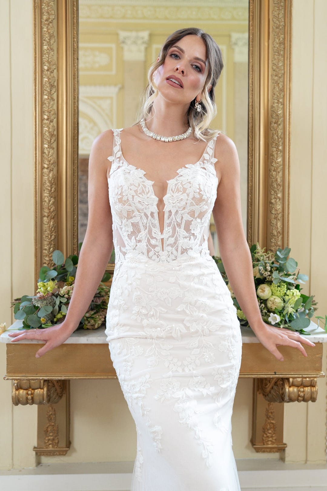 V26-F13_Web_2 Eine Frau in der Bridonista V26-F13 steht vor einem goldgerahmten Spiegel, hinter ihr Blumenarrangements auf der Konsole. Mit gewelltem Haar und einer Halskette blickt sie sanft in die Kamera und fängt zeitlose Eleganz ein.