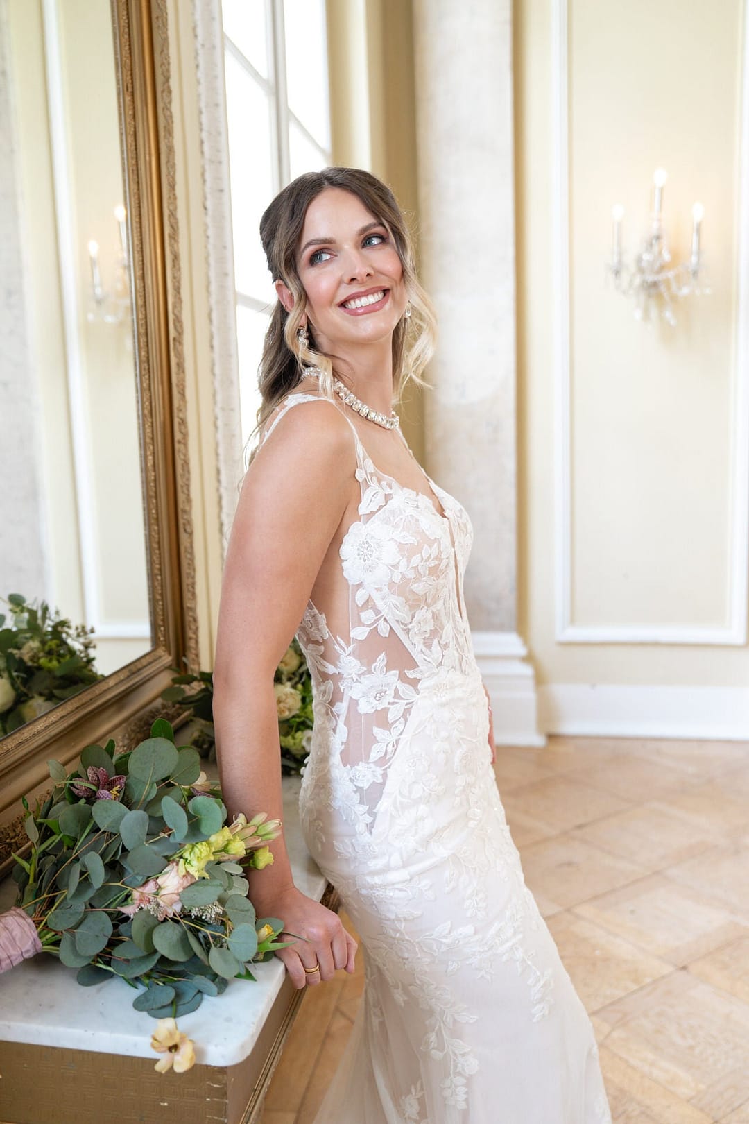 V26-F13_Web_3 Eine lächelnde Frau im ärmellosen Brautkleid aus Spitze von Bridonista V26-F13 steht in einem Innenraum vor einem Spiegel und einem Blumenstrauß, während das Sonnenlicht durch ein Fenster mit eleganter Dekoration im Hintergrund fällt.