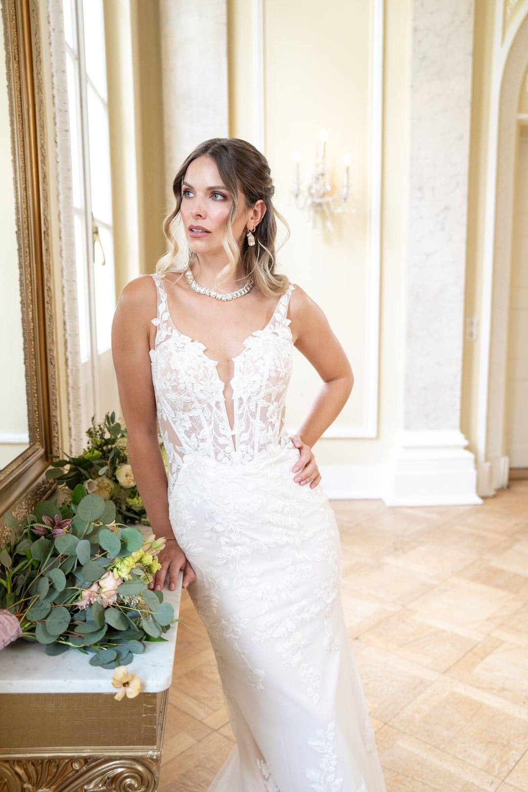 V26-F13_Web_4 Eine Frau in einem weißen, spitzenbesetzten, ärmellosen Bridonista V26-F13 Brautkleid steht in einem Innenraum neben einem mit Blumen geschmückten Marmortisch und schaut nachdenklich zur Seite. Sie hat gewelltes Haar und trägt eine Halskette und Ohrringe.