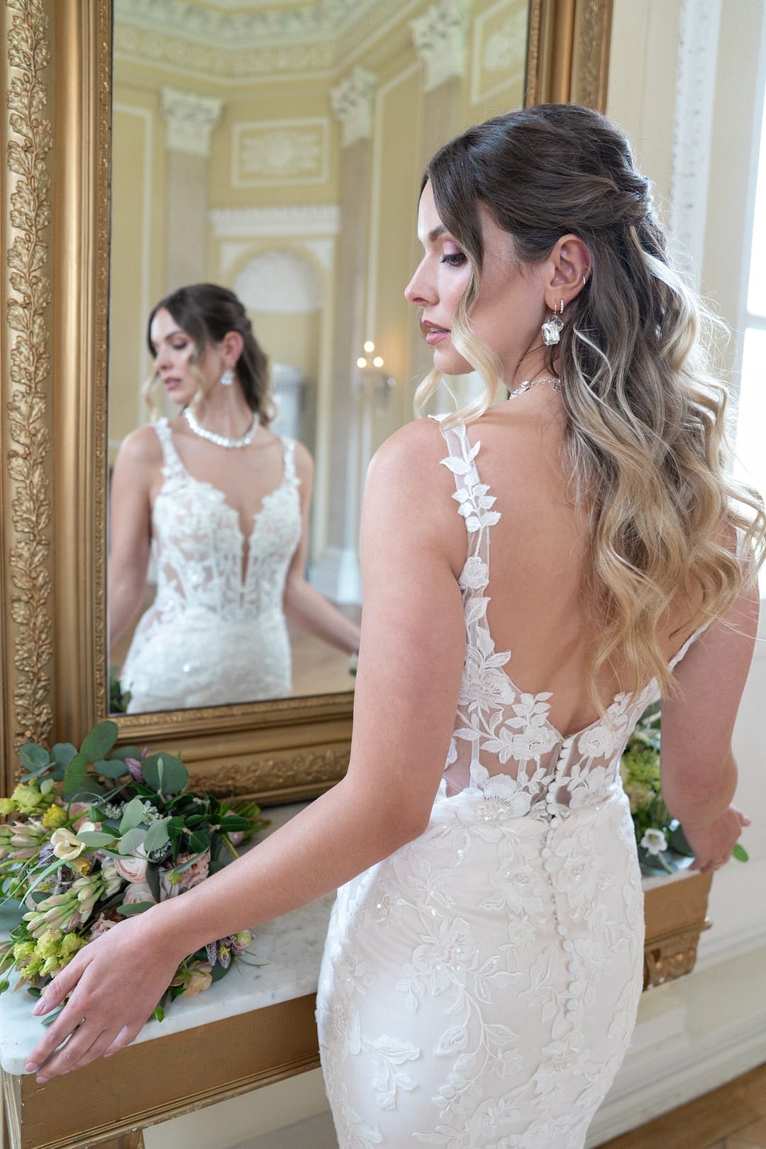 V26-F13_Web_5 Eine Braut mit gewelltem Ombre-Haar steht vor einem Spiegel in einem geschmückten Raum. Sie trägt das Bridonista V26-F13 Spitzen-Hochzeitskleid und hält einen Strauß grüner und weißer Blumen in der Hand; ihr Spiegelbild ist inmitten des klassischen Dekors zu sehen.