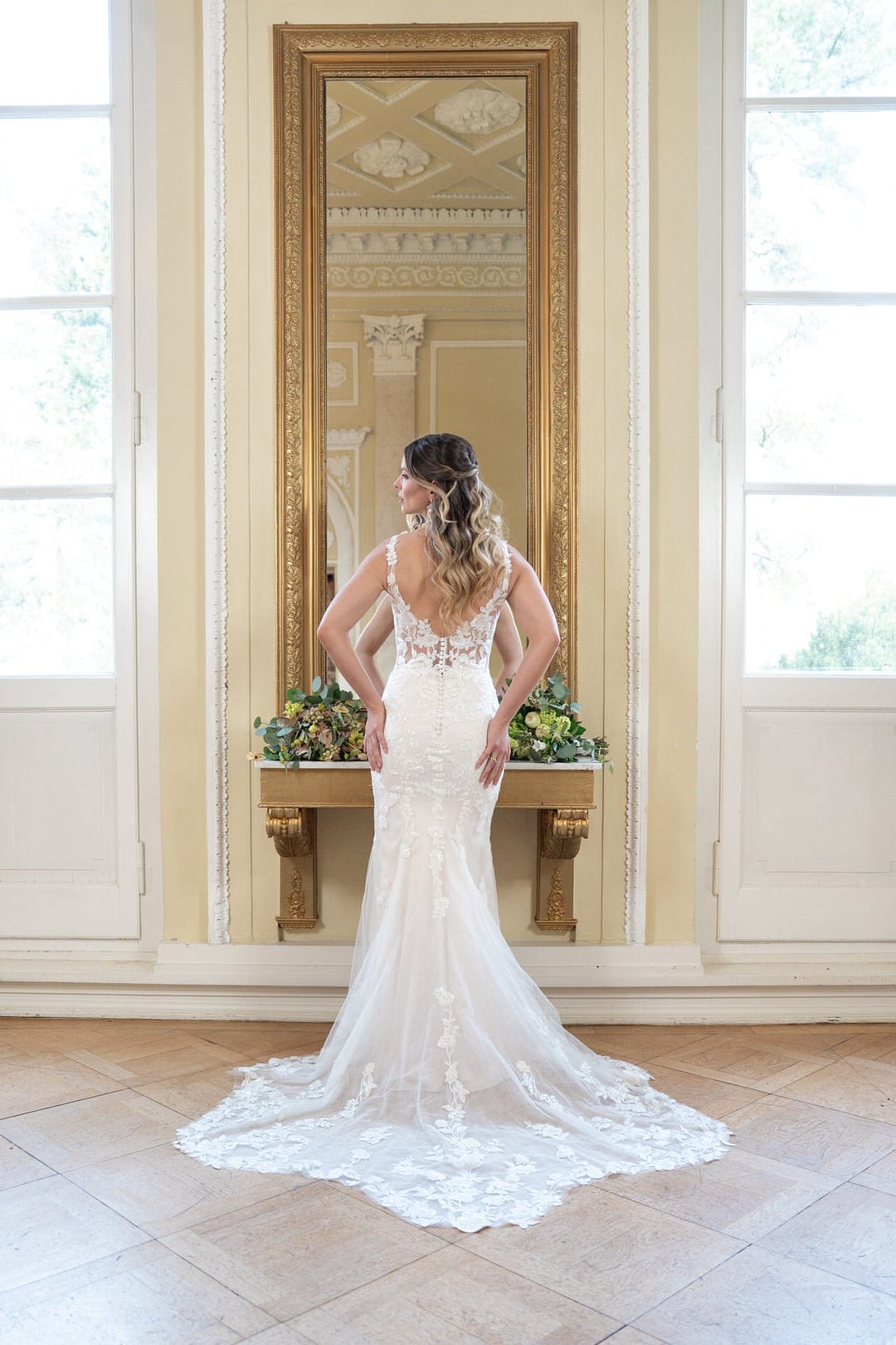 V26-F13_Web_6 Eine Braut, die das Bridonista V26-F13 trägt, steht vor einem hohen Spiegel in einem eleganten Raum mit großen Fenstern, die Hände in die Hüften gestemmt, das gewellte Haar fällt ihr in Kaskaden über den Rücken.