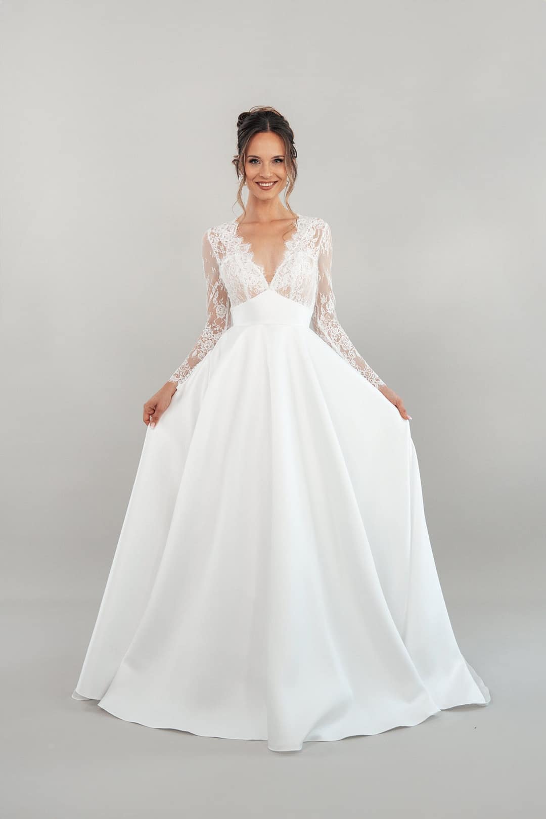Eine Frau steht lächelnd vor einem schlichten hellgrauen Hintergrund. Sie trägt das Produkt S26-B04 - ein langes weißes Hochzeitskleid mit Spitzenmieder und -ärmeln - und hält die Seiten ihres Rocks.