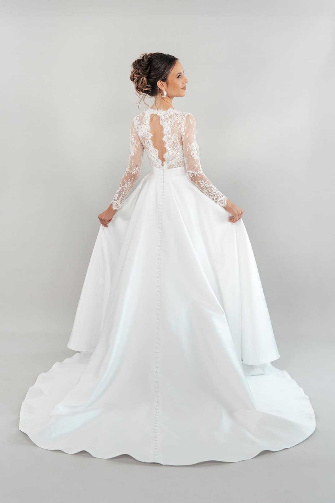 Eine Braut, die das lange weiße Hochzeitskleid S26-B04 mit Spitzenärmeln und tiefem Rücken trägt, steht vor einem schlichten hellen Hintergrund und hält die Seiten ihres Kleides.