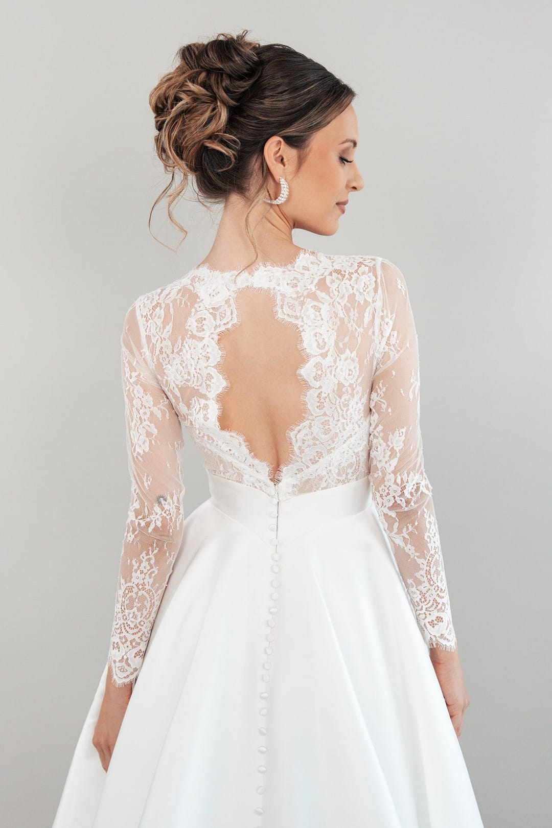 Eine Frau in dem Hochzeitskleid S26-B04 mit Spitzenärmeln und tiefem Rückenausschnitt steht mit einer eleganten Hochsteckfrisur vor einem schlichten hellgrauen Hintergrund.
