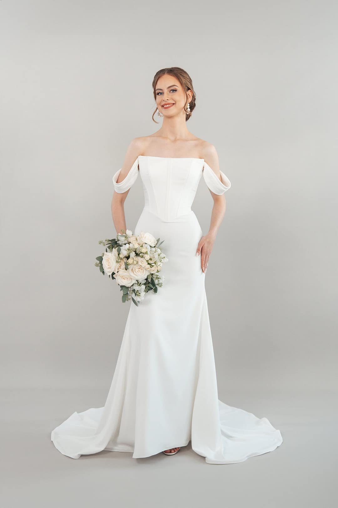 S26-F01_Web_1 Eine Frau, die das schulterfreie weiße Hochzeitskleid S26-F01 trägt, hält lächelnd einen Strauß weißer und blassrosa Blumen vor einem schlichten hellgrauen Hintergrund.