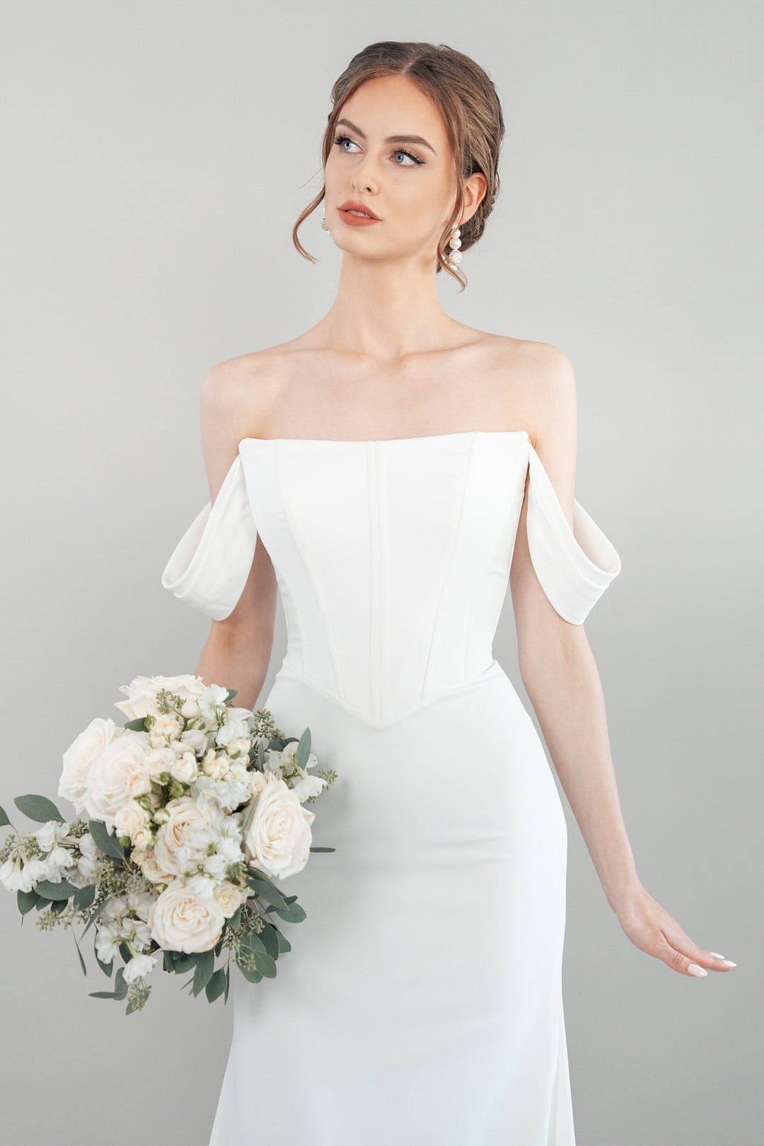 S26-F01_Web_3 Eine Frau modelliert das schulterfreie weiße Hochzeitskleid S26-F01 und hält einen Strauß weißer Blumen und Grünzeug vor einem schlichten hellgrauen Hintergrund. Ihr Haar ist hochgesteckt, während sie leicht nach oben blickt.