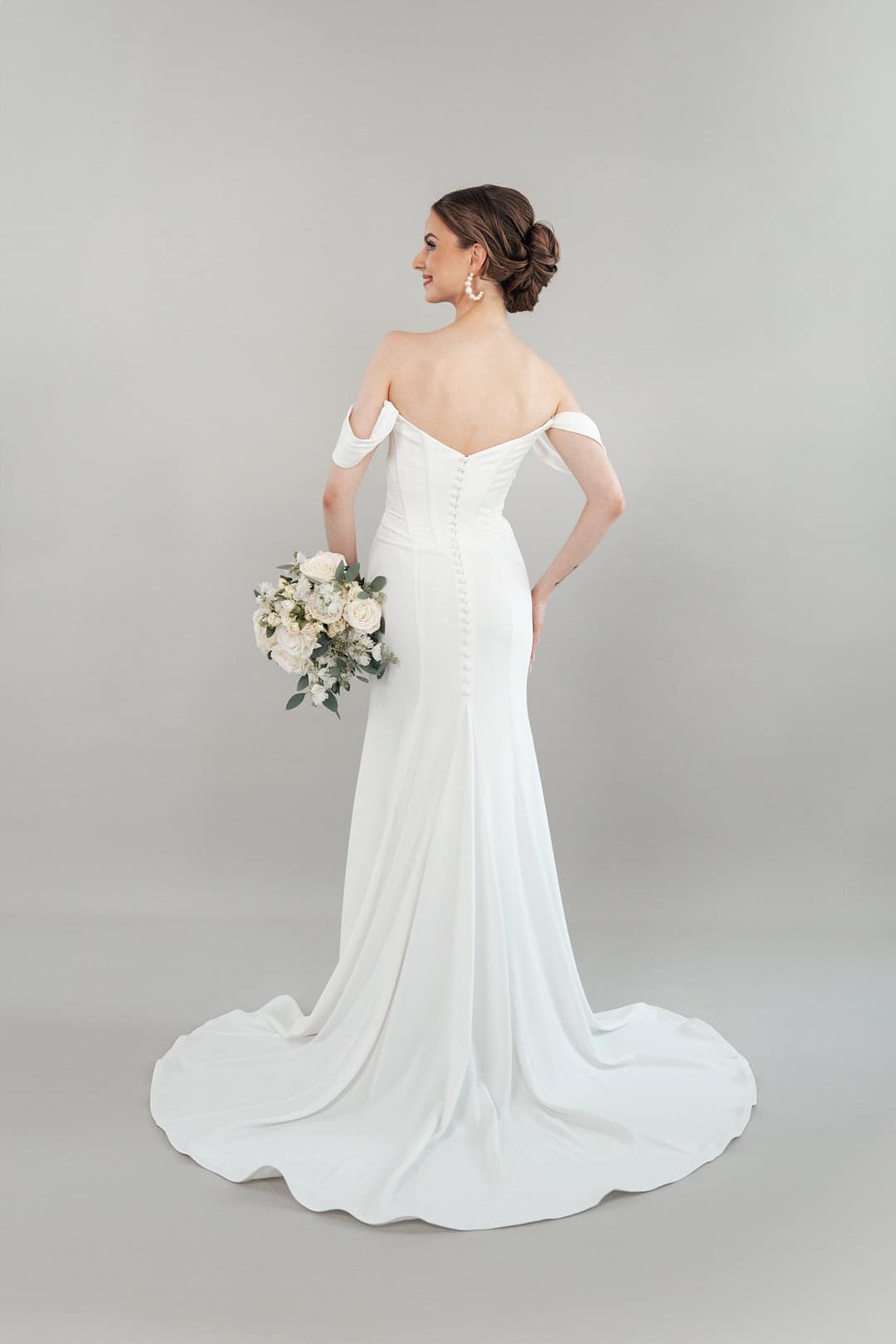 S26-F01_Web_4 Eine Braut im schulterfreien weißen Hochzeitskleid S26-F01 hält einen Strauß weißer Blumen und posiert mit dem Rücken, um den geknöpften Rücken und die Schleppe vor einem hellgrauen Hintergrund zu zeigen.
