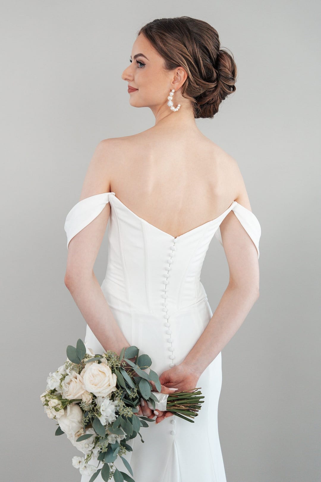 S26-F01_Web_5 Eine Frau, die das schulterfreie weiße Hochzeitskleid S26-F01 trägt, steht mit dem Rücken zugewandt und hält einen Strauß mit weißen Blumen und Grünzeug in der Hand. Ihr Haar ist elegant gestylt und sie trägt Perlenohrringe.