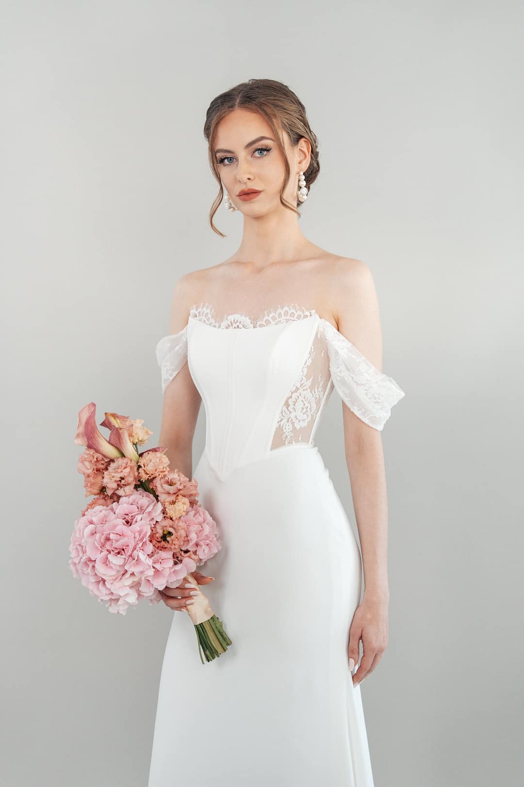 Eine Braut im schulterfreien weißen Hochzeitskleid S26-F09 mit Spitzendetails hält einen rosa Blumenstrauß. Ihr hellbraunes Haar ist hochgesteckt und sie trägt baumelnde Ohrringe. Sie posiert vor einem schlichten hellen Hintergrund.