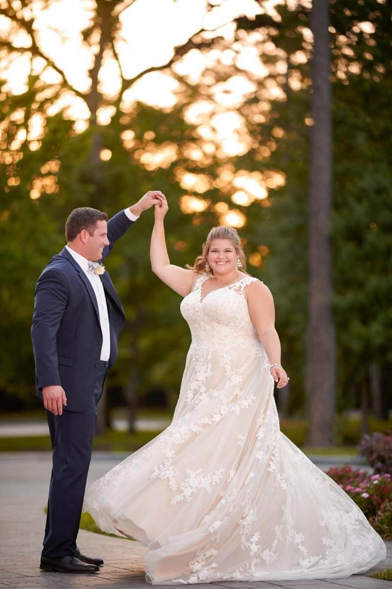 Eine Braut im weißen Spitzen-Brautkleid wirbelt lächelnd mit einem Bräutigam im dunklen Anzug und mit Fliege im Freien bei Sonnenuntergang umher, während sie sich an den Händen hält, mit Bäumen im Hintergrund. Die Szene fängt zeitlose Brautmoden-Eleganz ein.