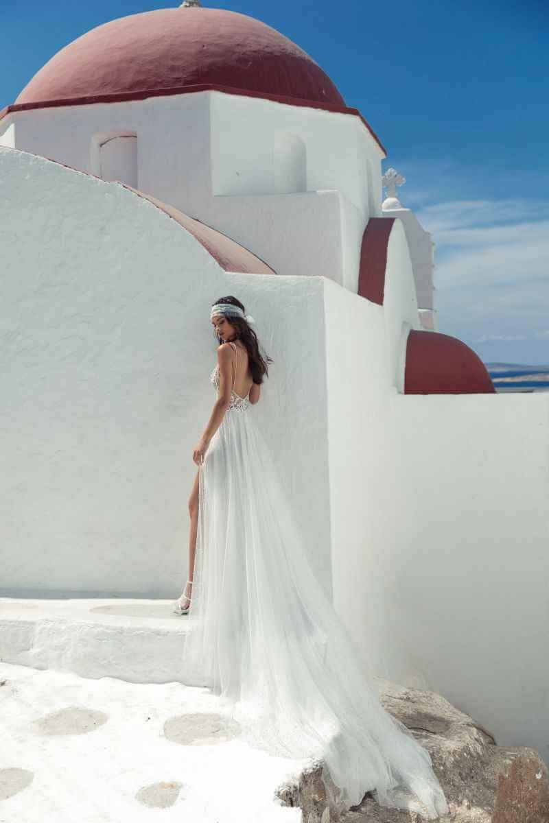 Eine Frau in einem fließenden weißen Brautkleid und Kopfstück steht auf weißen Steinstufen neben einem weiß getünchten Gebäude mit roter Kuppel, unter einem strahlend blauen Himmel.