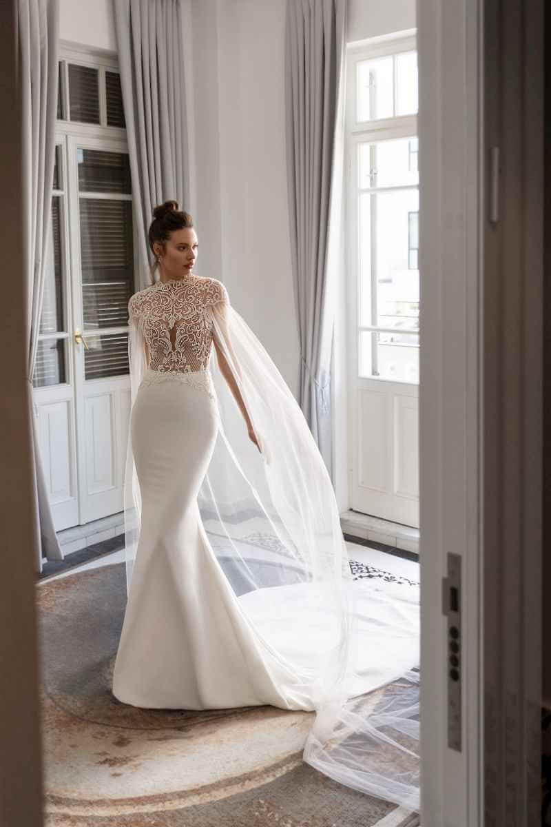 Eine Frau in einem taillierten, eleganten weißen Brautkleid mit Spitzenoberteil und transparentem Cape steht in einem hellen, großzügigen Raum mit großen Fenstern und hellgrauen Vorhängen - ein Traum aus moderner Brautmode.
