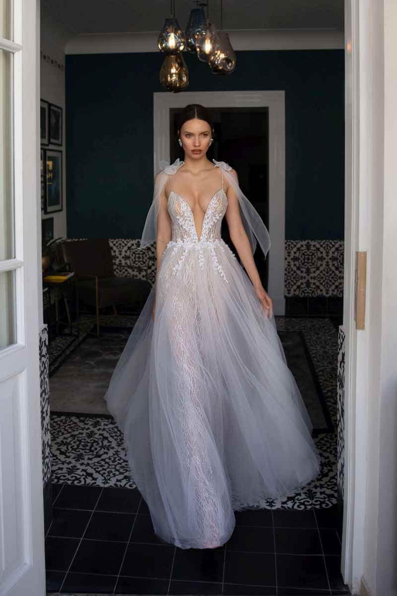 Eine Frau steht in einem Innenraum und trägt ein weißes Hochzeitskleid aus Spitze mit tiefem Ausschnitt, durchsichtigem Tüllüberzug und Schleifenverzierung. Sie wird von einer offenen Tür eingerahmt, hinter der sich ein dunkles, elegantes Interieur befindet.