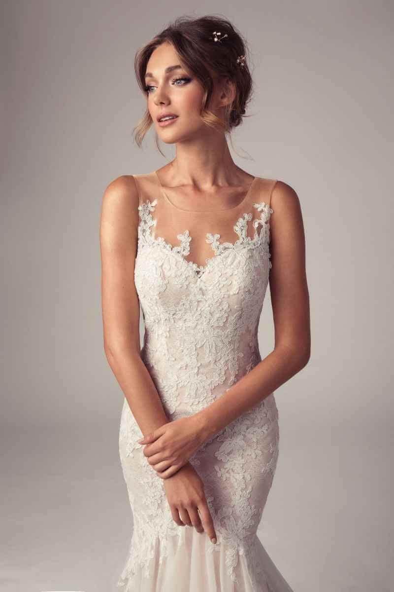 Eine Frau, die ein tailliertes Hochzeitskleid aus Spitze mit durchsichtigen Trägern und Blumenapplikationen trägt, steht vor einem neutralen Hintergrund und blickt zur Seite, während sie ihre Hände sanft in der Taille verschränkt.