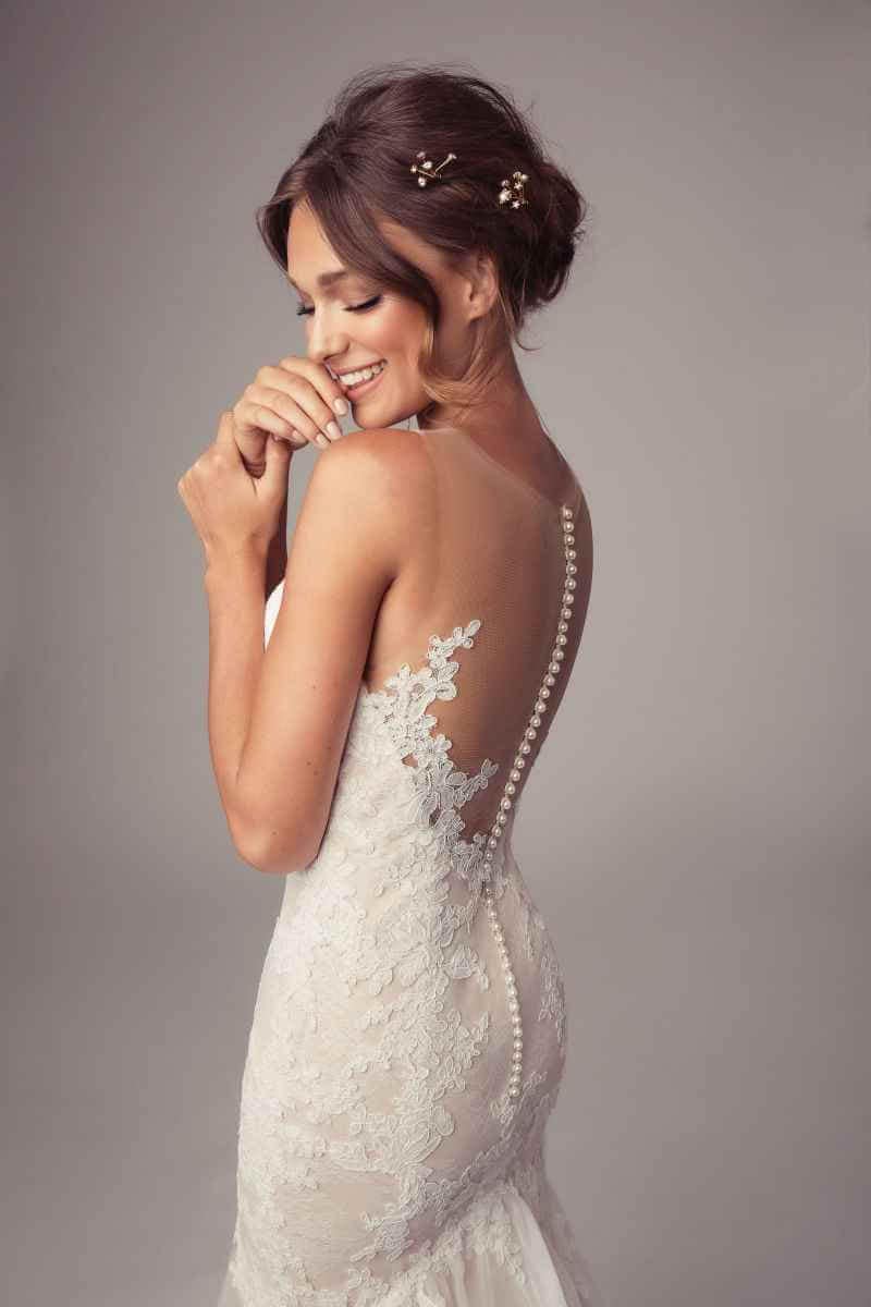 Eine lächelnde Braut in einem spitzenbesetzten Hochzeitskleid mit durchsichtigem Rücken und dekorativen Knöpfen steht im Profil, ihr Haar in einer Hochsteckfrisur mit kleinen floralen Akzenten, vor einem neutralen Hintergrund.
