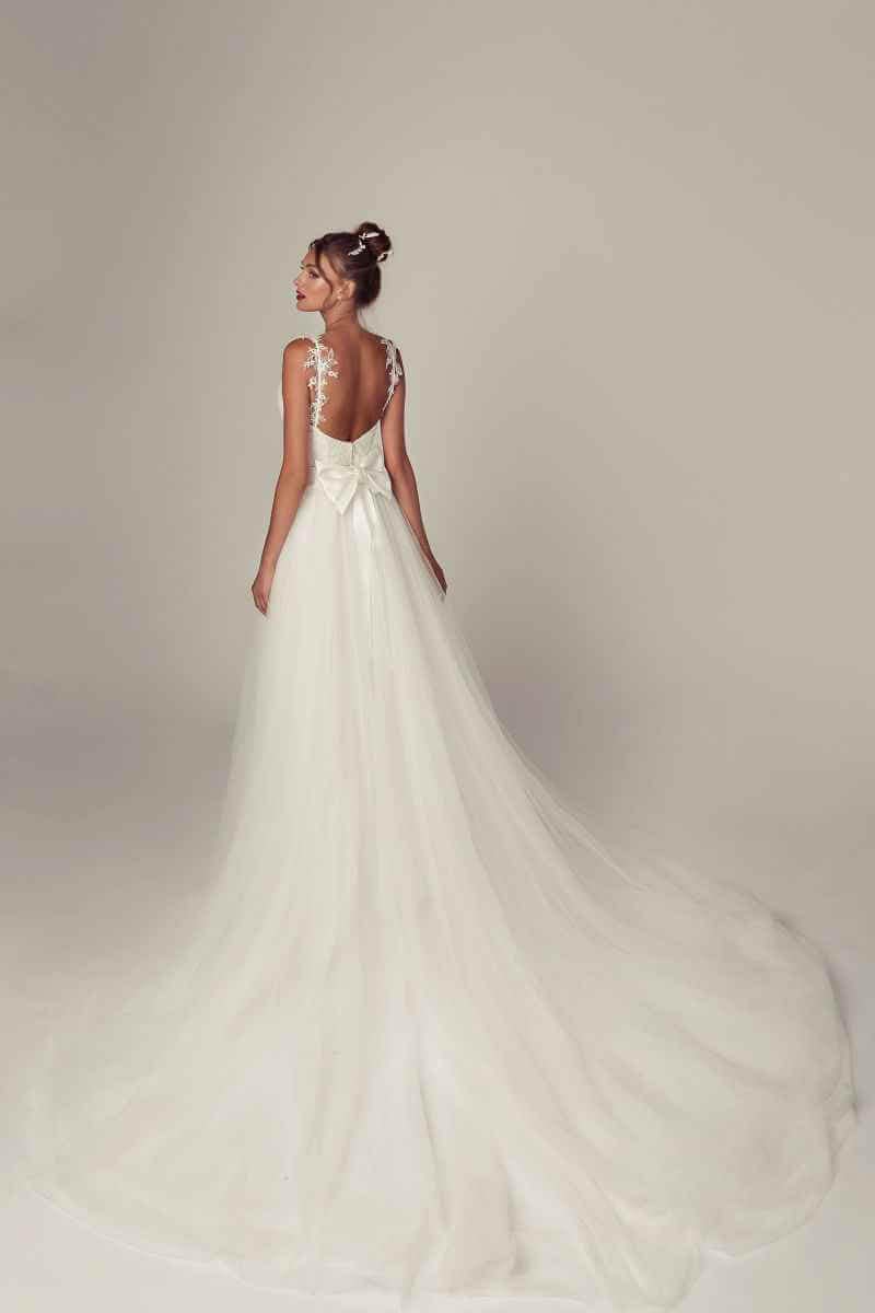 Eine Frau in einem langen, fließenden weißen Hochzeitskleid mit Spitzenträgern und einer großen Schleife auf dem Rücken steht mit dem Rücken zugewandt und blickt über ihre Schulter vor einem schlichten, hellen Hintergrund.