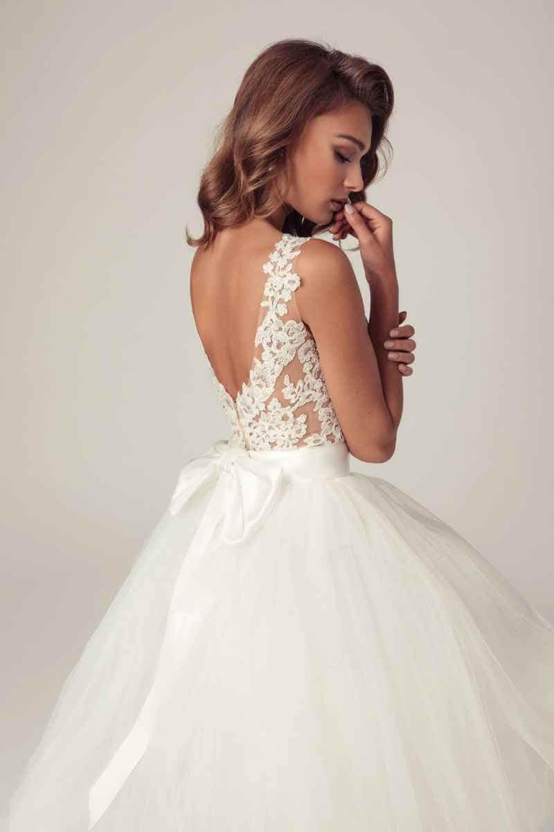 Eine Frau in einem weißen Brautkleid mit Spitzenrücken, Tüllrock und Schleife in der Taille steht im Profil und blickt nachdenklich mit einer Hand am Gesicht nach unten - eine zeitlose Wahl von eleganten Brautmoden.