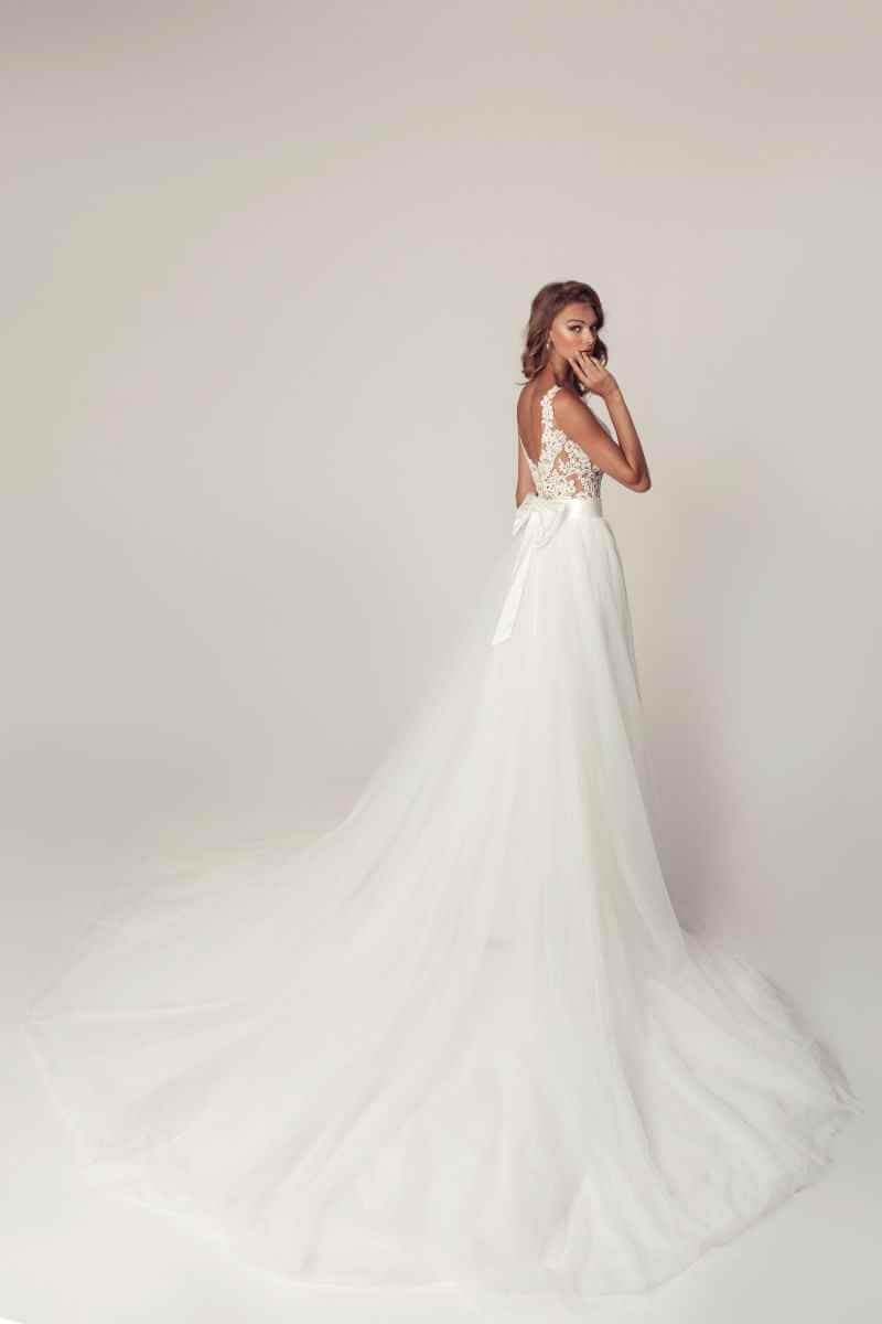 Eine Frau in einem langen, eleganten Hochzeitskleid mit Spitzenrücken und fließender Schleppe steht leicht zur Seite gedreht in einem gedämpft beleuchteten Atelier und schaut über ihre Schulter.