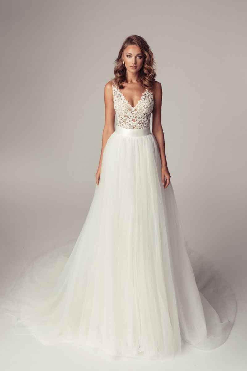 Eine Frau, die ein weißes Hochzeitskleid mit einem Spitzenmieder und einem vollen Tüllrock trägt, steht vor einem neutralen Hintergrund und blickt nach vorne.