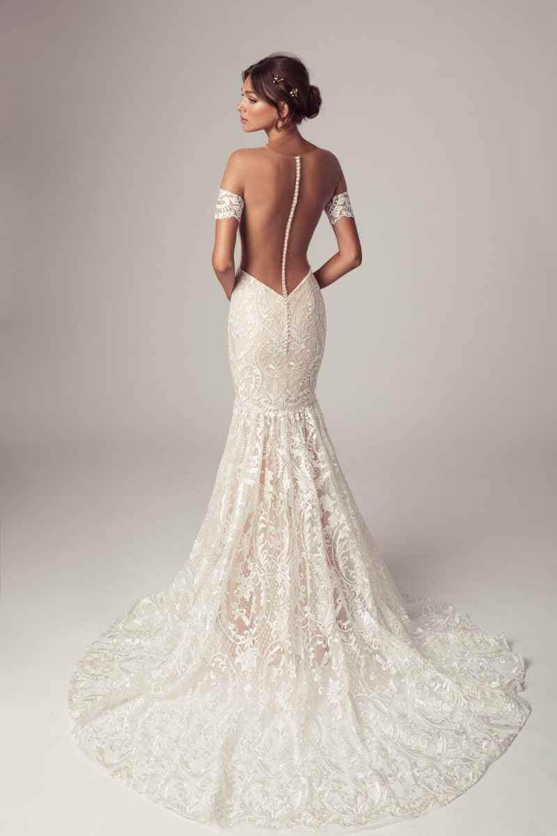 Eine Frau in einem eleganten, schulterfreien Spitzen-Hochzeitskleid mit dramatisch offenem Rücken und Knopfleiste steht mit dem Gesicht zur Wand, das Haar zu einem tiefen Dutt frisiert und mit einem dezenten Haarschmuck versehen.