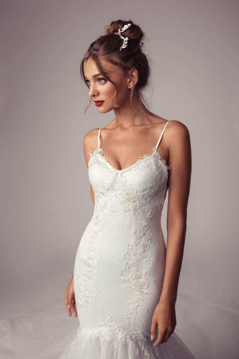 Eine Frau in einem taillierten, weißen Spitzenbrautkleid mit dünnen Trägern steht vor einem neutralen Hintergrund. Ihr Haar ist zu einer eleganten Hochsteckfrisur mit dekorativen Haarnadeln gestylt, ergänzt durch roten Lippenstift und dezentes Make-up.