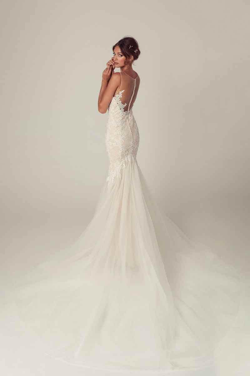 Eine Frau steht mit dem Blick über die Schulter in einem taillierten, rückenfreien weißen Brautkleid mit Spitzenstickerei und einer langen, fließenden Tüllschleppe. Der Hintergrund ist einfarbig und hell.