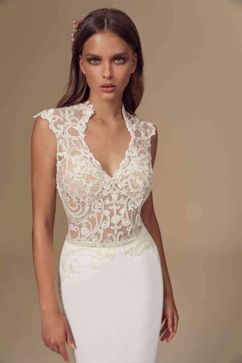 Eine Frau in einem ärmellosen, mit Spitze bestickten Hochzeitskleid mit tiefem V-Ausschnitt steht vor einem schlichten beigen Hintergrund. Sie hat langes braunes Haar und einen neutralen Gesichtsausdruck.