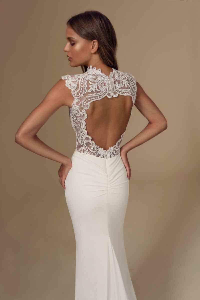 Eine Frau trägt ein elegantes weißes Brautkleid mit taillierter Silhouette, aufwändiger Spitzenverzierung am oberen Rücken und offener Rückenpartie. Sie steht vor einem neutralen Hintergrund und blickt von der Kamera weg.