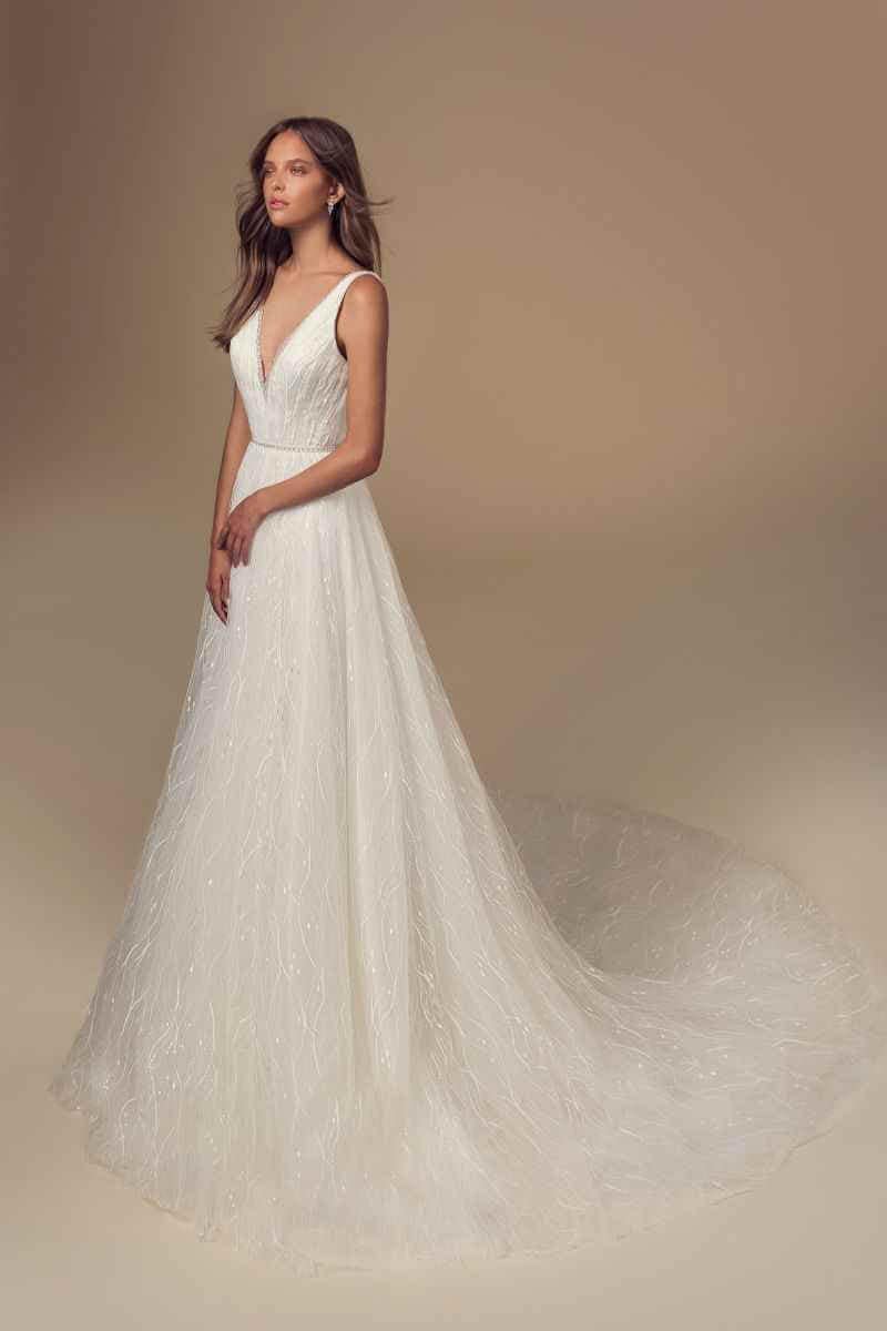 Eine Frau, die ein ärmelloses, bodenlanges weißes Brautkleid mit tiefem V-Ausschnitt und langer, fließender Schleppe trägt, steht vor einem beigen Hintergrund.