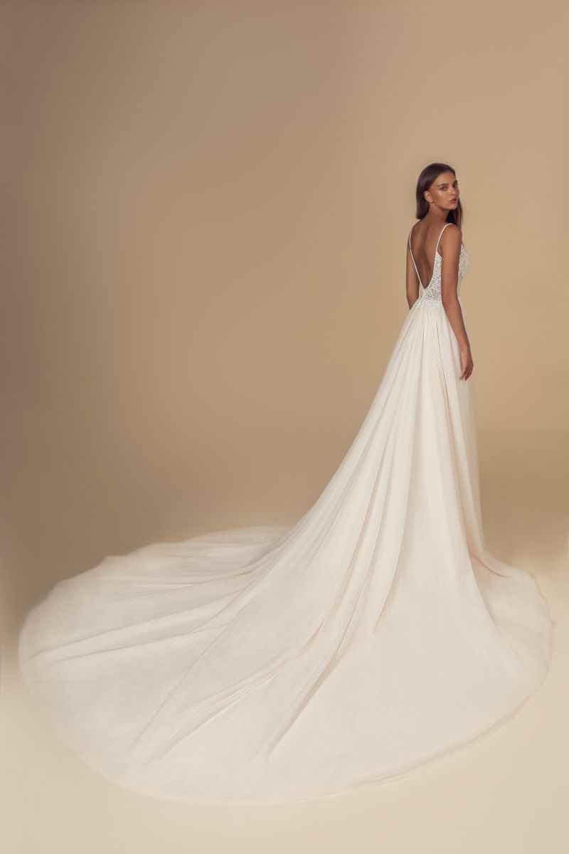 Eine Frau in einem weißen Hochzeitskleid mit einer langen, fließenden Schleppe steht vor einem beigen Hintergrund und blickt über ihre Schulter. Das Brautkleid hat einen tiefen V-Rücken und zarte Träger.