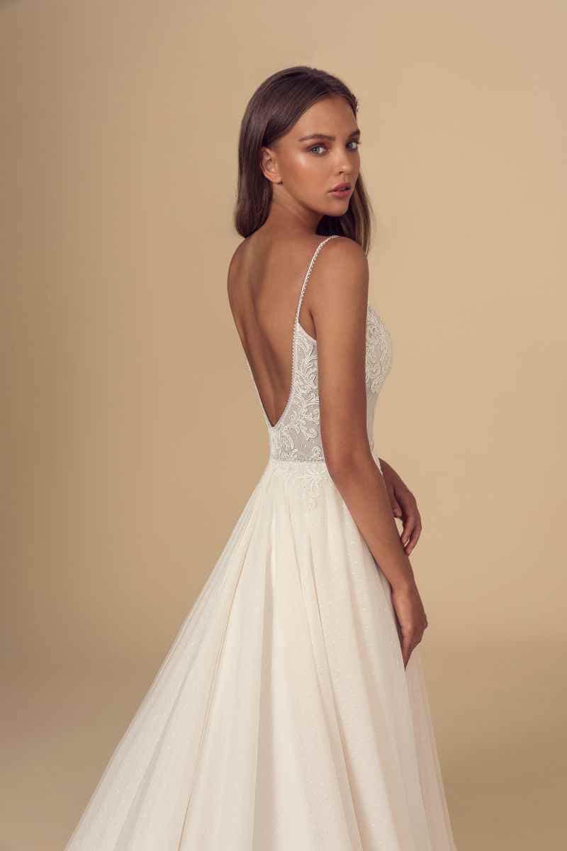 Eine Frau in einem ärmellosen, weißen Hochzeitskleid mit Spitzendetails und tiefem V-Rücken steht seitlich und schaut über ihre Schulter vor einem einfarbig beigen Hintergrund.